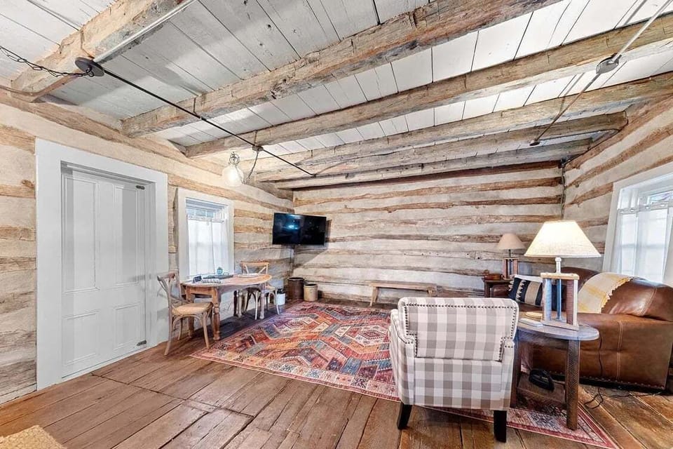 The living area provides a leather sofa, plaid armchair, wall-mounted television, dining table, and warm historic finishes.