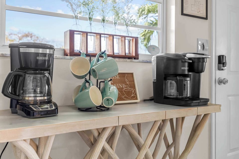 Coffee station with two coffee makers and a set of mugs on a light wood table by the window