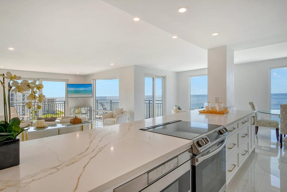 Marble kitchen island with built-in cooktop overlooks bright living area and ocean-facing windows.