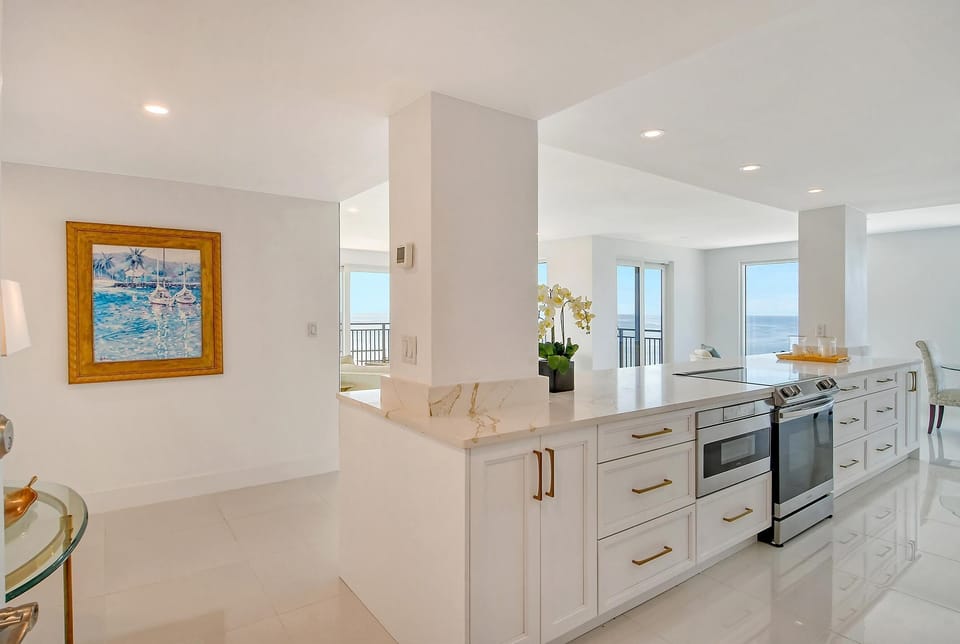 Marble-topped kitchen island with brass handles and stainless steel appliances next to ocean-facing windows.