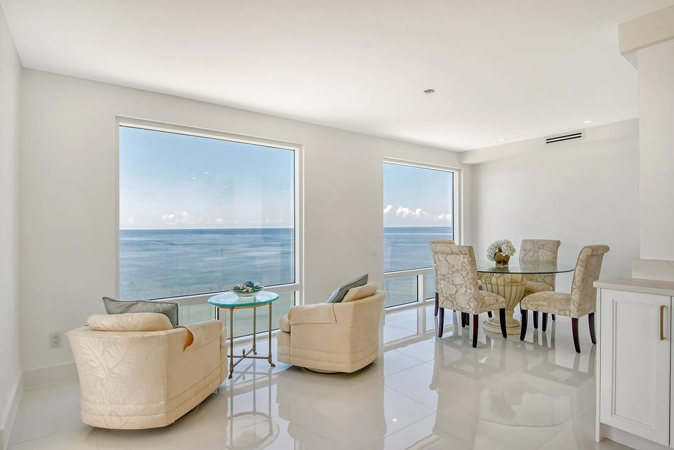 Dining area with round glass table, four patterned chairs, and beige armchairs facing expansive ocean windows.