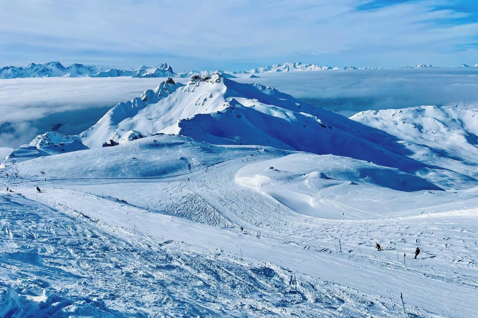 Wide open snow-covered slopes with tiny skiers dotting the vast white terrain under a bright blue sky.