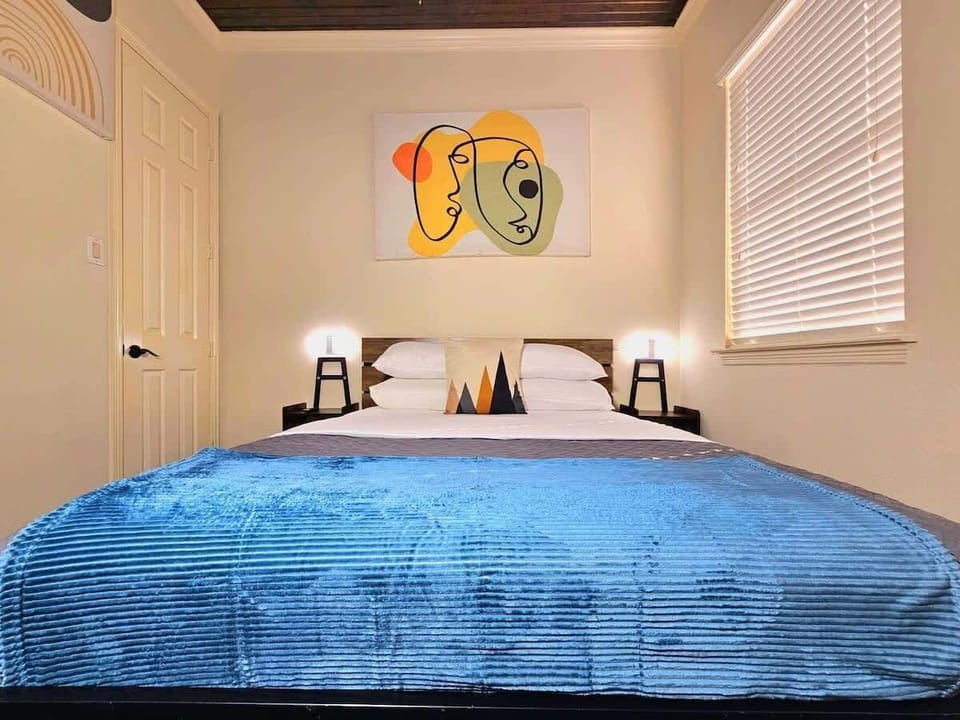 Cozy bedroom with blue blanket, abstract wall art, and warm bedside lamps.

