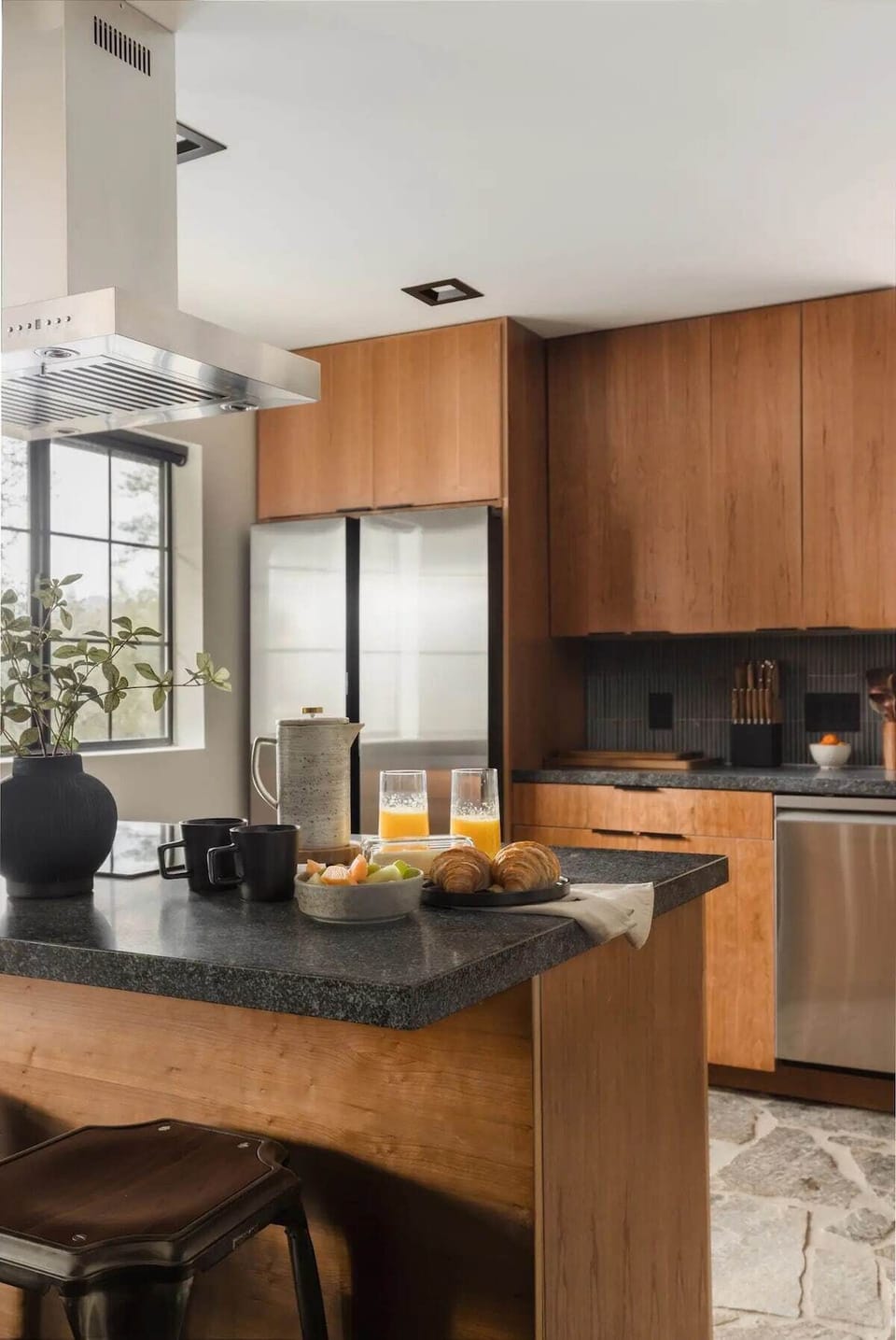 A modern, warm kitchen, featuring sleek wood cabinetry and a spacious island with comfortable stools. The kitchen is equipped with stainless steel appliances, including a large, modern refrigerator.