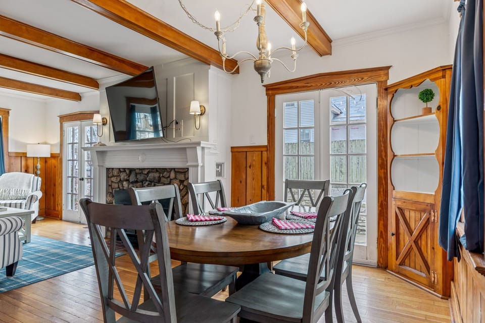 A round dining table beneath a chandelier sits between the living room, kitchen, and stone fireplace.