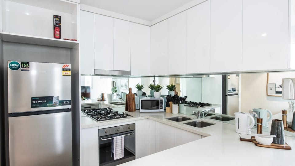 A pristine, wrap-around white kitchen featuring premium integrated appliances and mirror splashbacks that amplify the space.