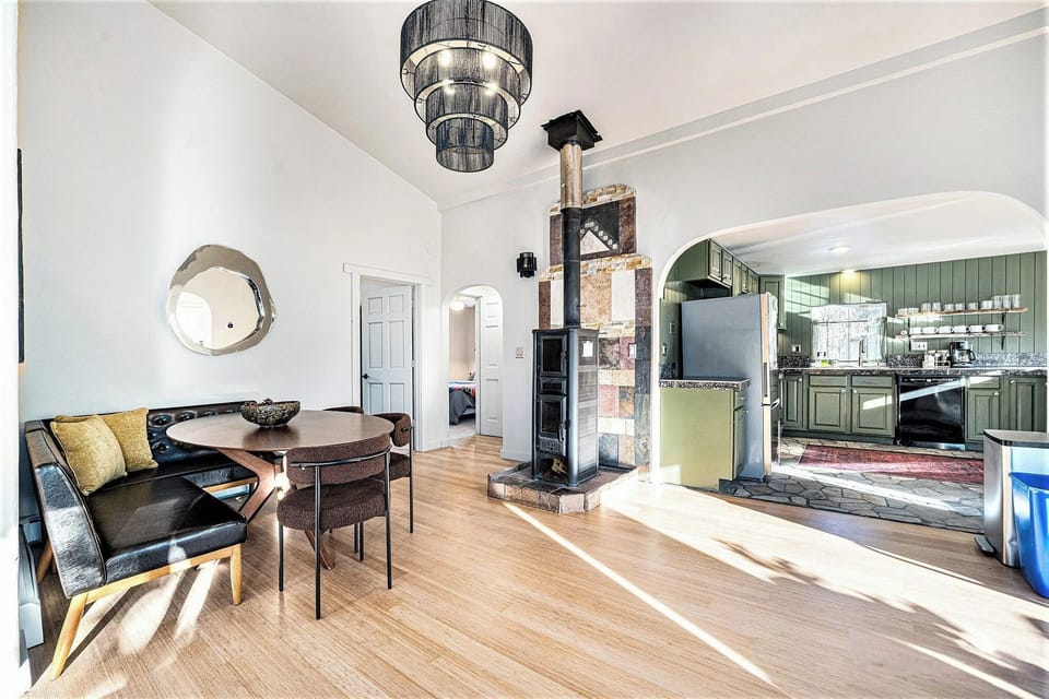 - Dine in a modern nook with tufted seating, sculptural chairs, and sunlit ambiance
- Unique tiled wood stove adds character and warmth to the open layout
- Olive green kitchen cabinets, floating shelves, and stone floors exude rustic elegance