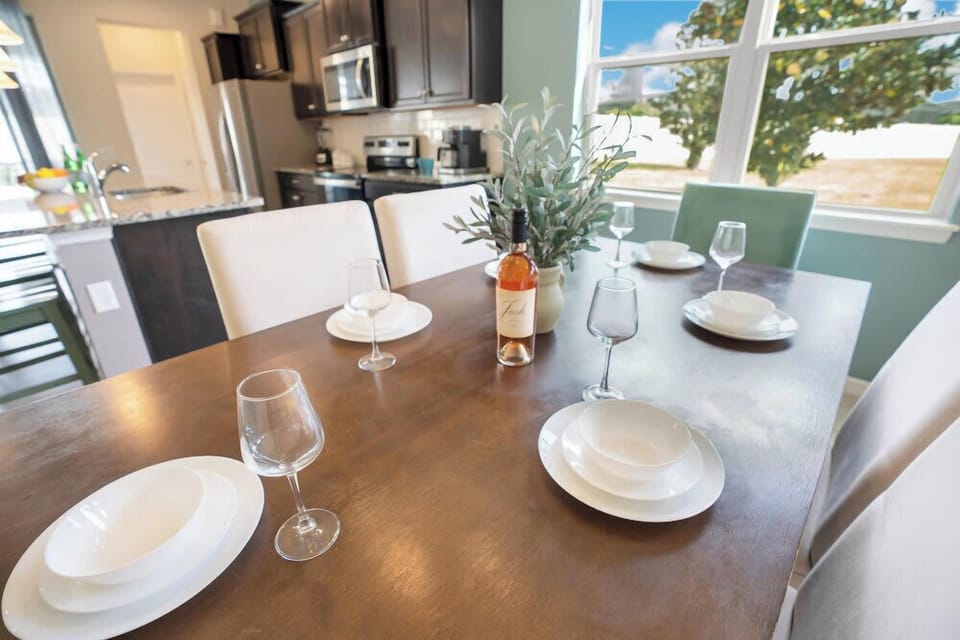 Dining area with a wooden table set for six and a view of the kitchen and window.