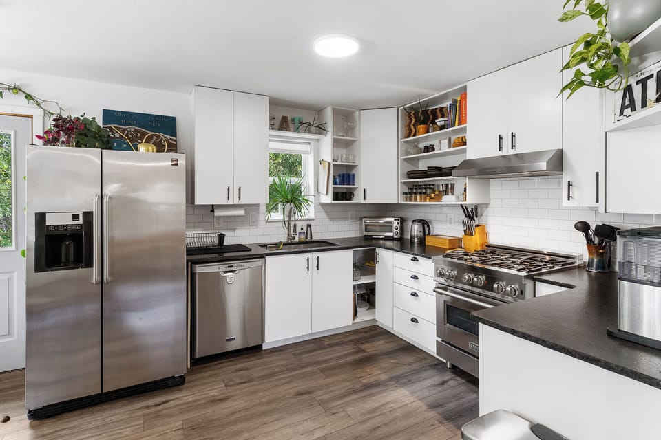 Modern kitchen with stainless steel appliances, white cabinets, a gas stove, double-door refrigerator, and wooden flooring. Potted plants and various kitchen items are visible on the countertops and shelves.