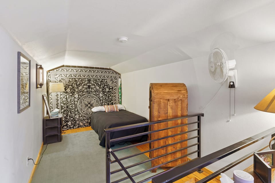 A small bedroom with a single bed, black and white patterned wall tapestry, wooden chest, nightstand with a lamp, and a wall-mounted fan.