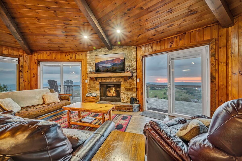 Spacious living room with vaulted ceilings, stone fireplace, and floor-to-ceiling windows showcasing stunning mountain views. The perfect spot for family movie nights or cozy cabin vibes.