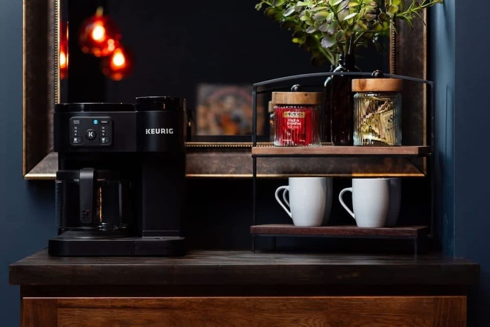 A neatly arranged coffee nook featuring a sleek Keurig machine, stylish mugs, and glass jars of coffee essentials, all complemented by warm lighting and a decorative mirror backdrop.
