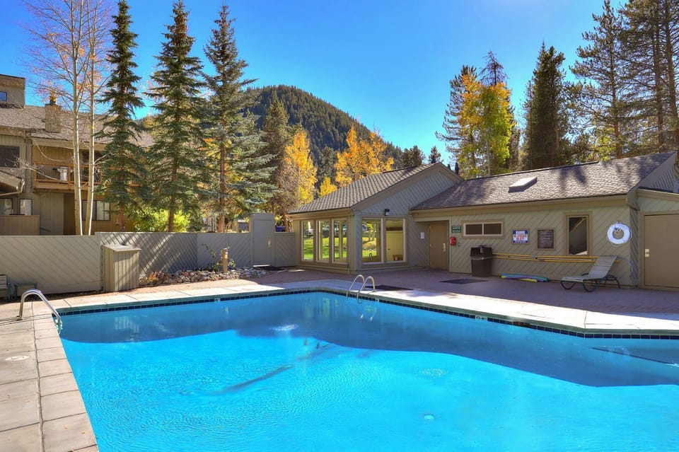 Outdoor swimming pool with clear blue water, surrounded by trees and a building with large windows. A lifebuoy is mounted on the wall, and mountains are visible in the background.