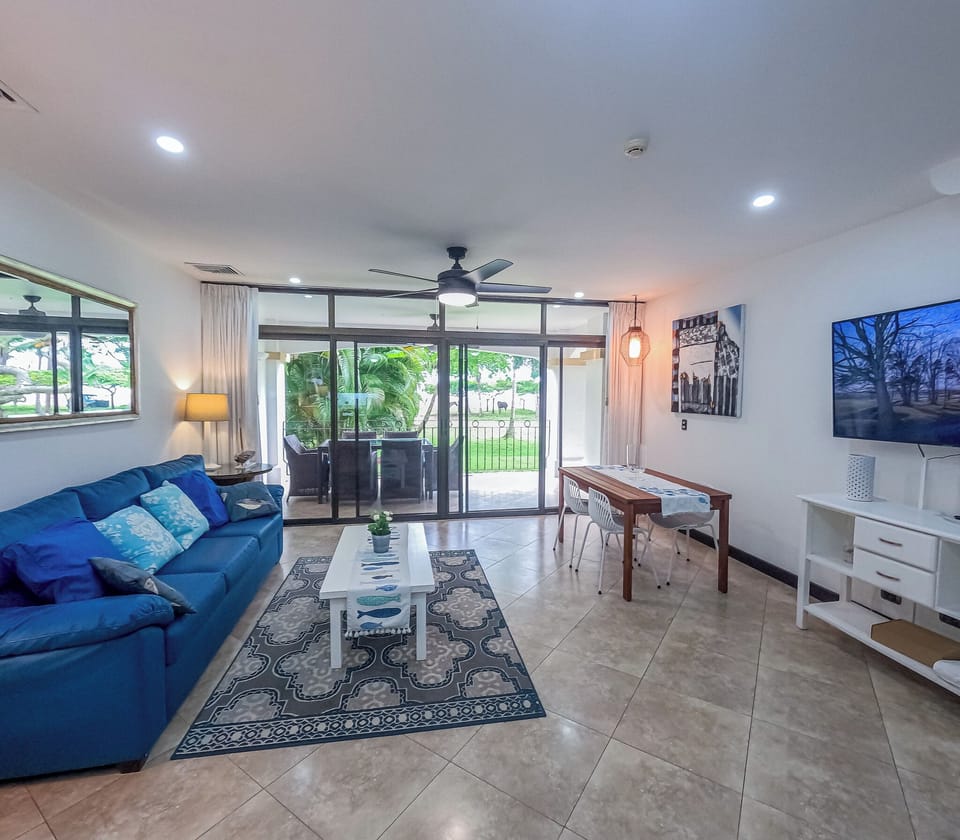 A spacious living room with a comfortable sofa, a smart TV, and a dining table perfect for your breakfast, lunch, and dinner, all with a beautiful view of the property's lush green area.