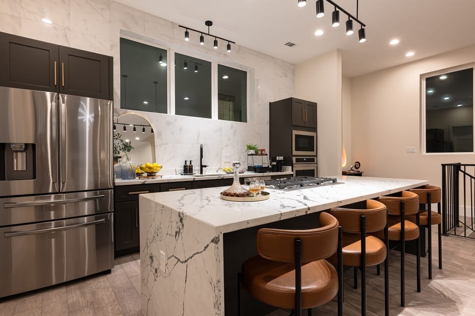 A modern kitchen with a large island, a stainless steel refrigerator, and dark cabinets. The kitchen has a large window with a view of the city. 