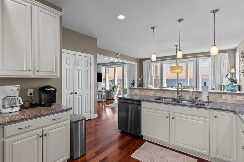 Bright coastal kitchen with white cabinets, double wall ovens, granite counters, stainless appliances, pendant lights, hardwood floors, and ocean views beyond the sink.