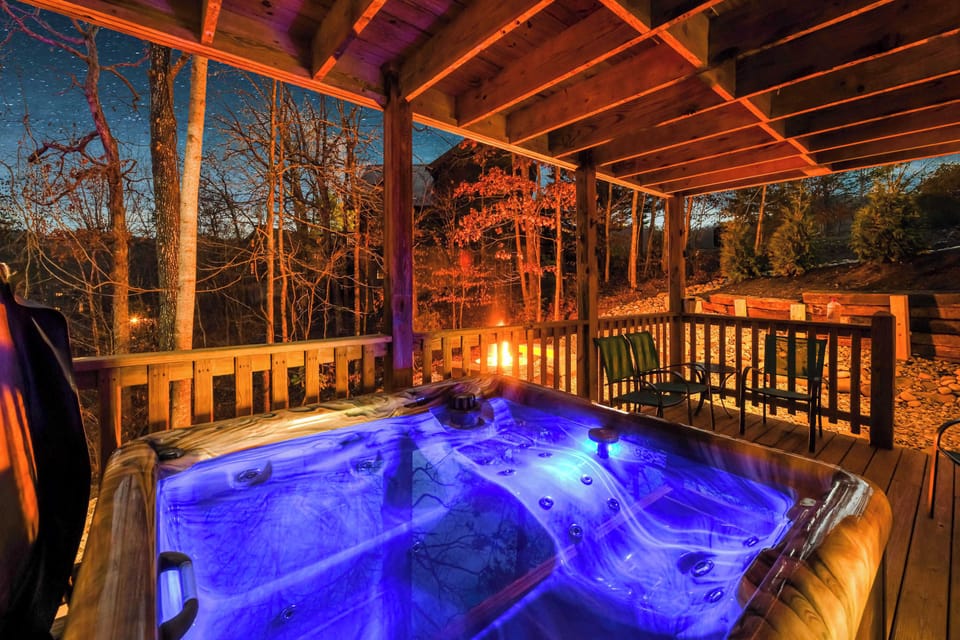 Relax in the illuminated hot tub on the covered wooden deck with a view of the forest and fire pit at night.