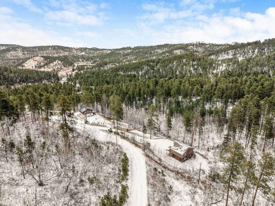 The aerial view showcases a house blanketed in snow, surrounded by trees, with a breathtaking winter landscape and distant rolling hills or mountains in the background.