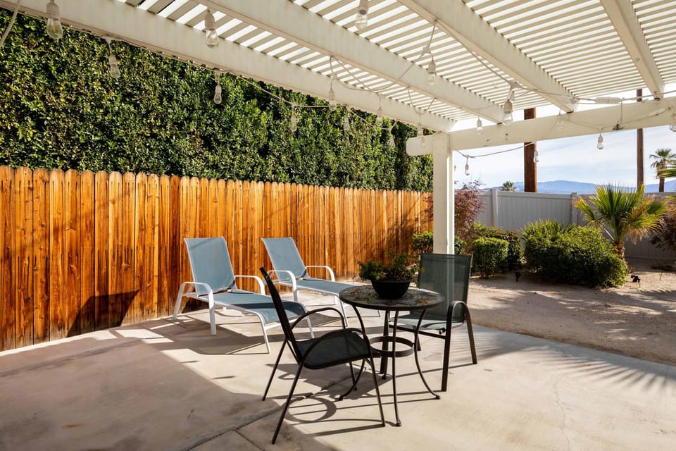Casita-side patio with pergola, string lights, sunlit loungers, dining set, and lush green privacy wall — a sheltered retreat of your own.