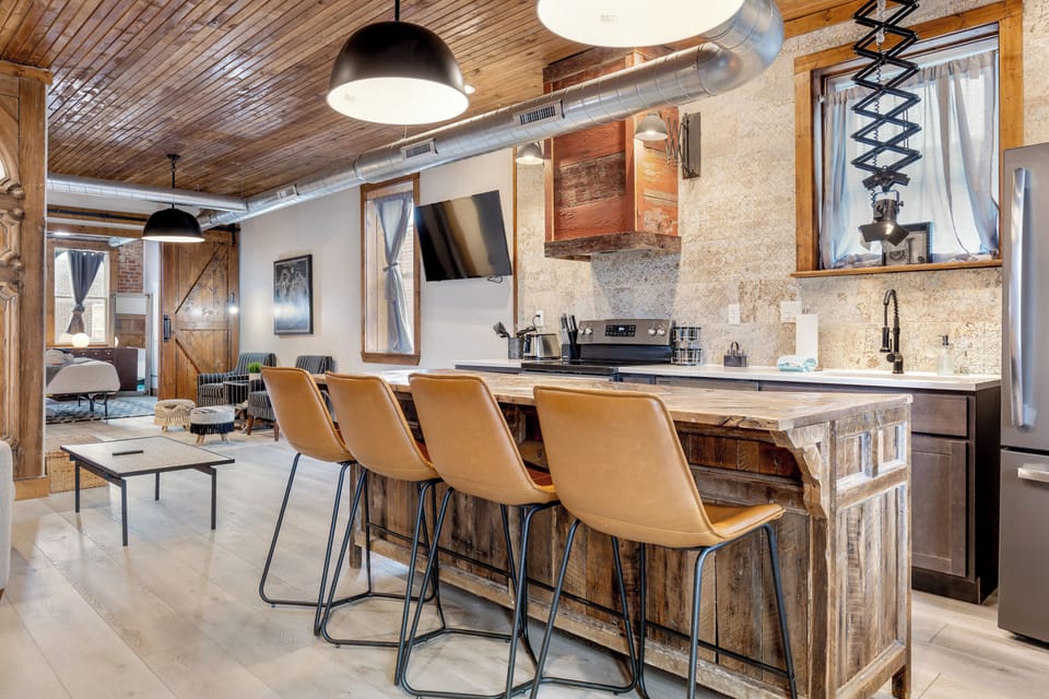 Four leather stools line the large kitchen island with the sitting area and bedroom visible beyond.