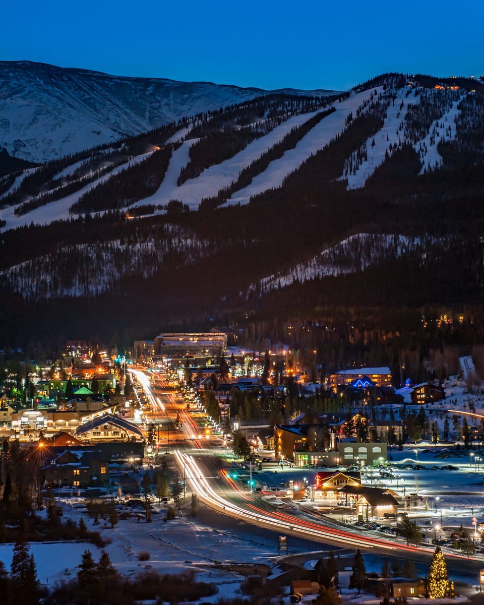 A stunning alpine ski resort glows warmly against snow-covered mountain slopes as evening lights illuminate the village streets and runs under a deep blue twilight sky.