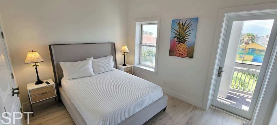 Airy coastal bedroom with soft white linens, warm wood floors, and balcony access overlooking palms. A bright, calming retreat designed for slow mornings and breezy evenings.