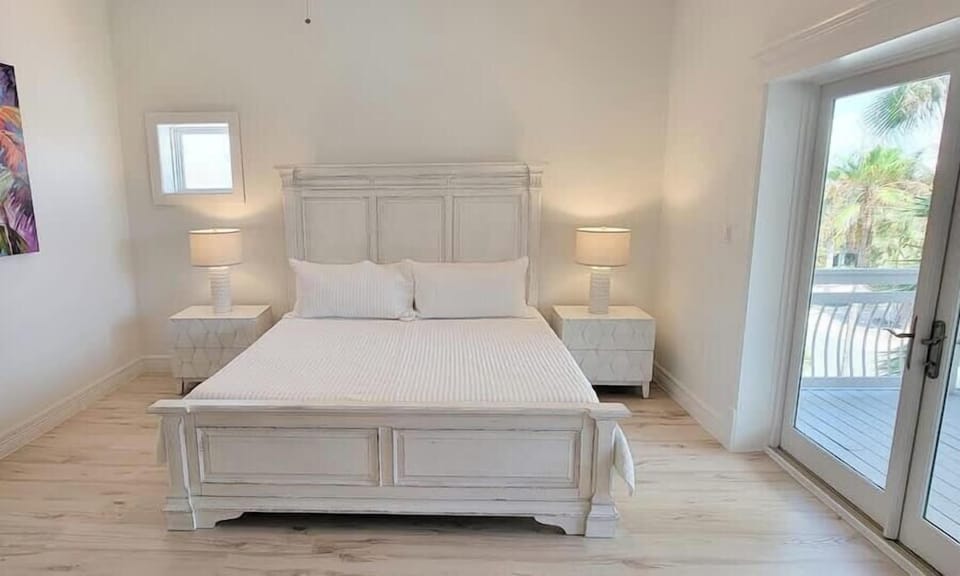 Bright coastal bedroom with crisp white décor, plush bedding, and private balcony access. Sun‑filled space with serene artwork, warm wood floors, and relaxing tropical views—perfect for unwinding after a beach day.