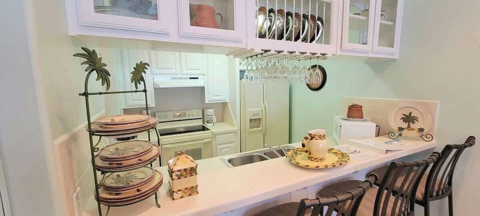 Open, welcoming dining space with a glass‑top table, woven chairs, a spacious breakfast bar overlooking a bright, organized kitchen. Warm lighting, greenery, and thoughtful décor create an easy, comfortable setting for shared meals and gatherings.
