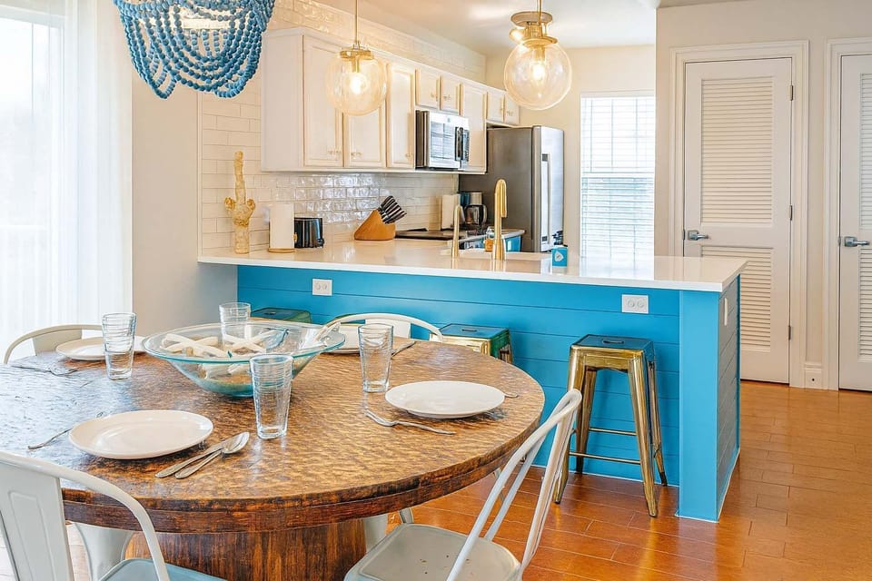 Bright and stylish dining space with a rustic round table for four, chic turquoise accents, and a modern kitchen with stainless appliances and breakfast bar seating—ideal for relaxing meals and gathering with friends or family.