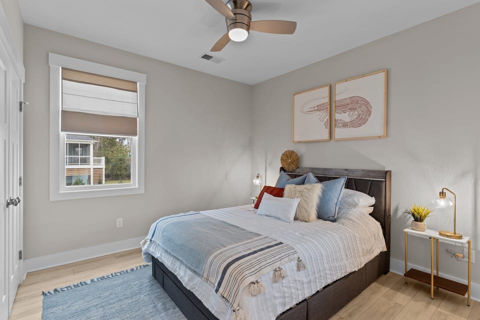 Coastal bedroom with queen bed, ceiling fan, window, woven shade, blue rug, soft pillows, side table lamps, wood floors, and shrimp wall art.