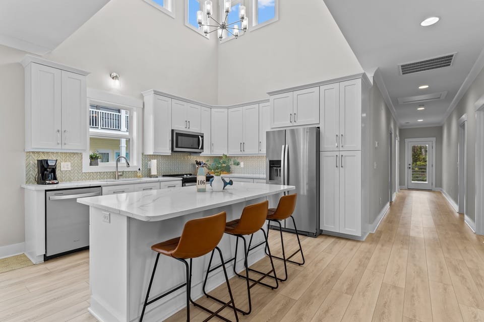 Bright coastal kitchen with vaulted ceilings, white cabinets, marble island, three stools, stainless appliances, tile backsplash, wood floors, and hallway entry.