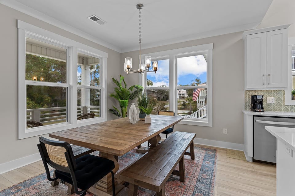 Bright dining space with wood table, bench, black chairs, chandelier, plants, large windows, porch views, patterned rug, and nearby modern kitchen.