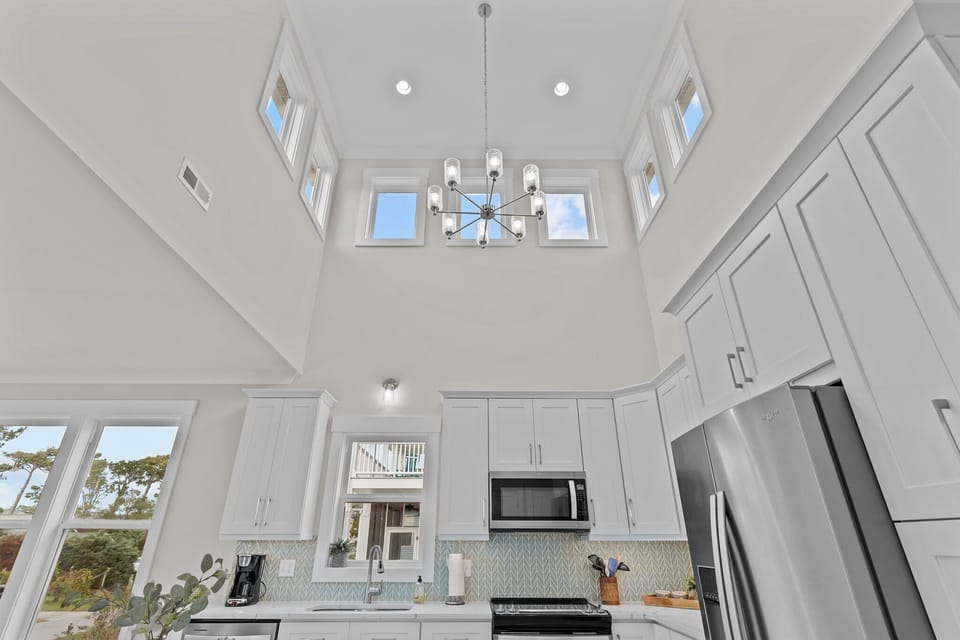 Bright coastal kitchen with vaulted ceilings, white cabinets, marble island, three stools, stainless appliances, tile backsplash, wood floors, and hallway entry.