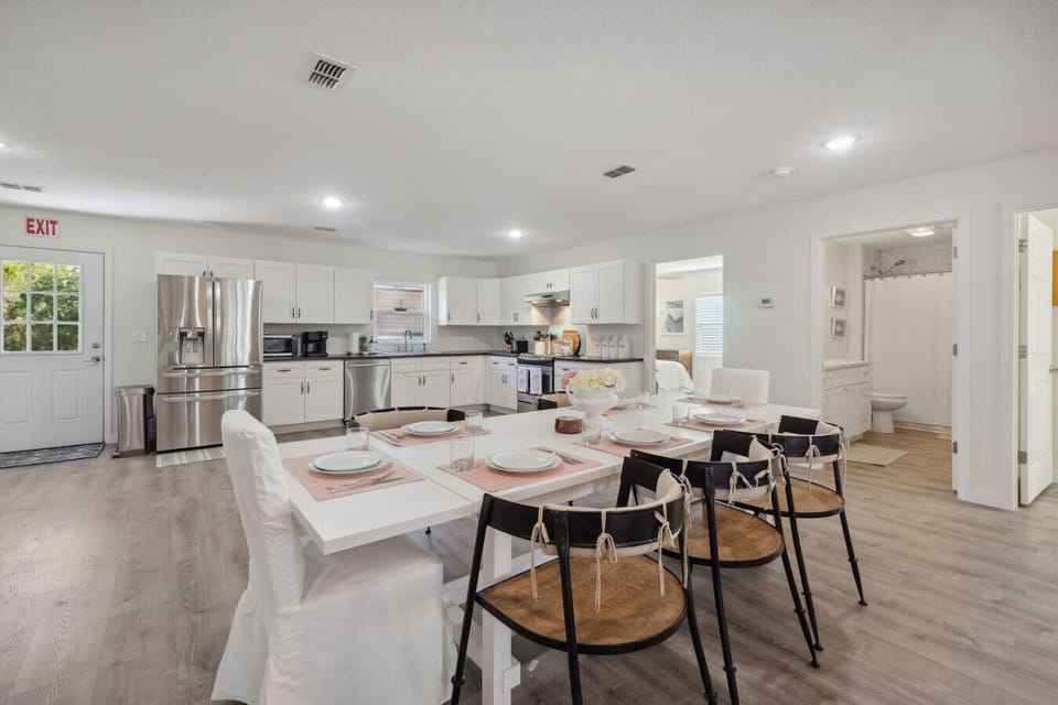 This bright and spacious open-concept kitchen and dining area featuring sleek white cabinetry, stainless steel appliances, and a large dining table set for seven. The clean design and natural light create a welcoming and functional space.
