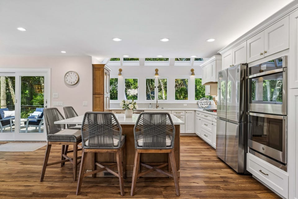 Floor-to-ceiling windows flood the room with natural light, offering serene views of lush greenery, a glimpse of the ocean, the private pool and spa, and the expansive deck.