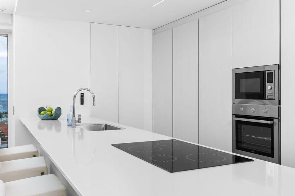 Your sleek, modern kitchen features stainless steel appliances, a glass cooktop, and a pristine white island—perfect for preparing meals while enjoying glimpses of blue water through the window.