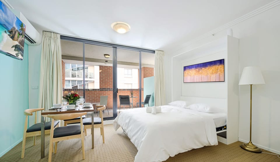 Bright studio layout showing the pull-up bed near the balcony, small dining table, and access to the private balcony.