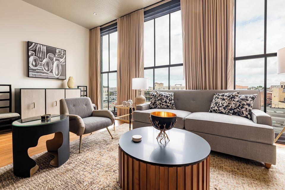 Inviting living space with large windows and natural light. The soft tones, modern furniture, and exposed brick create a comfortable and warm atmosphere.