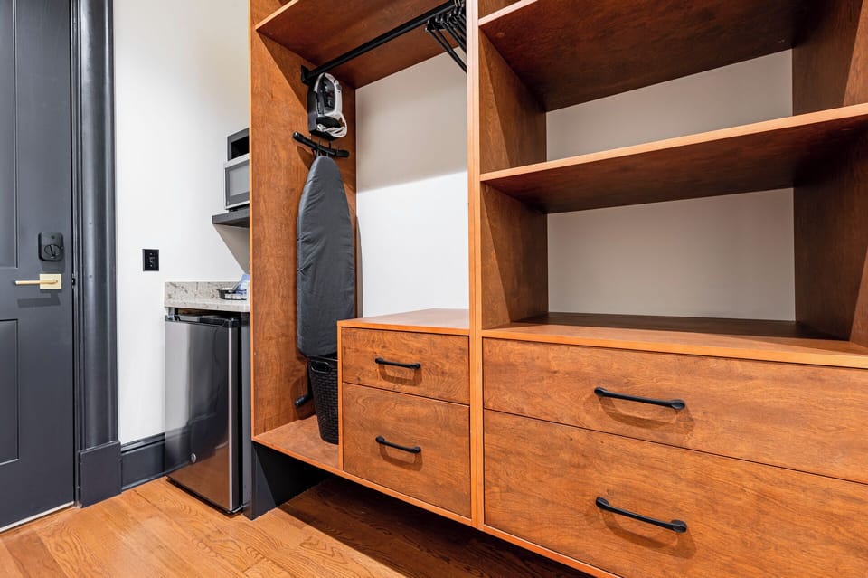 Closet space equipped with hangers, laundry hamper, iron with ironing board, coffee station, mini-fridge, and microwave.