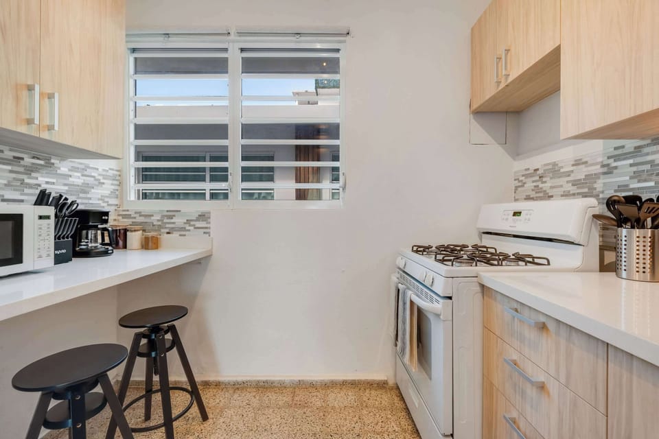 A narrow galley kitchen with light wood cabinets, a stainless steel gas stove, oven, and refrigerator. White countertops line both sides, with framed artwork on the wall and a wooden runner rug on the floor.