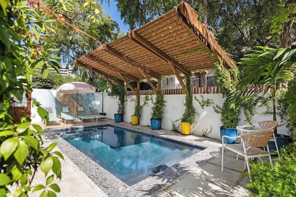 An outdoor pool surrounded by tropical plants, with a wooden pergola providing shade over lounge chairs. The pool is lined with blue tiles, and greenery surrounds the seating area.
