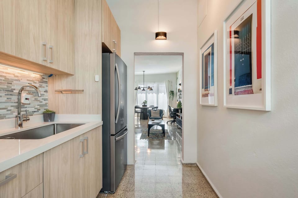 A narrow galley kitchen with light wood cabinets, a stainless steel gas stove, oven, and refrigerator. White countertops line both sides, with framed artwork on the wall and a wooden runner rug on the floor.