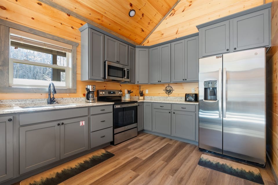 Fully equipped kitchen with gray cabinetry, stainless appliances, and a warm wood ceiling—newer build feel with cabin character. Cook real meals, not just microwave survival food. Coffee, breakfast, and dinner are easy here.
