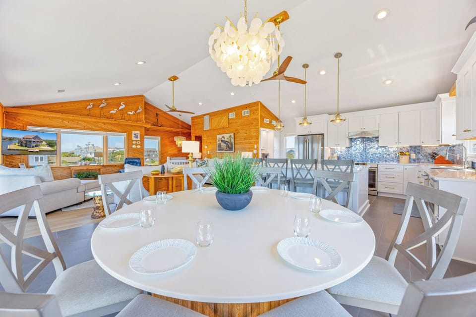 An open dining and kitchen area features soaring ceilings, elegant lighting, and a stunning backsplash that adds coastal charm to your group gatherings.