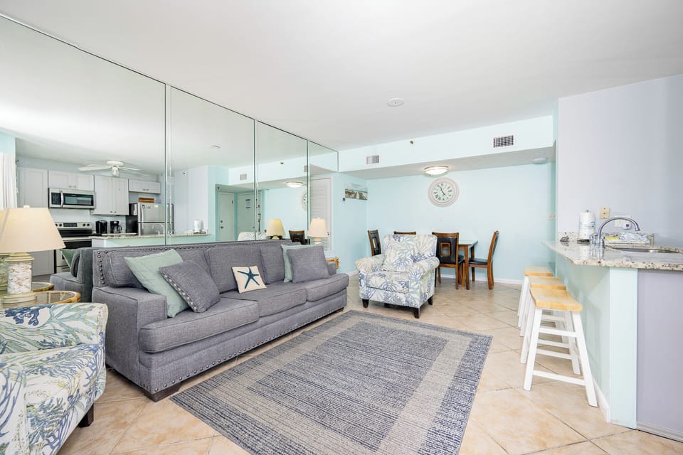The living space features a cozy gray sofa, vibrant accent chairs, and a view of the well-equipped kitchen and dining area