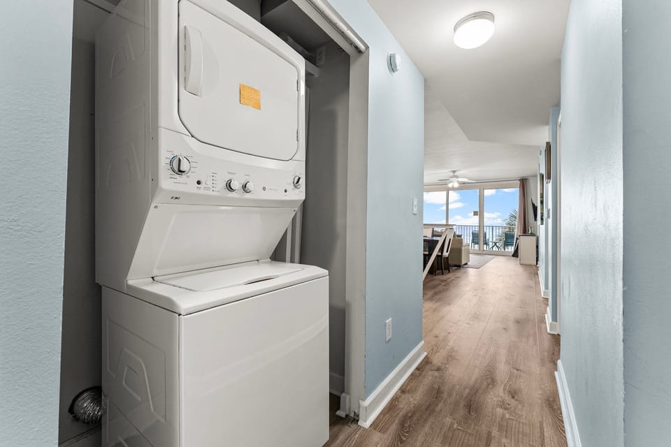 Stacked washer and dryer in a modern hallway leading to a bright living area with ocean views.