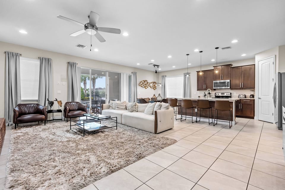 Enjoy a bright and spacious living area featuring a cozy light sofa and rich brown leather chairs. The warm wooden kitchen cabinetry contrasts with sleek tile flooring, and large windows bring in natural light and lovely views.