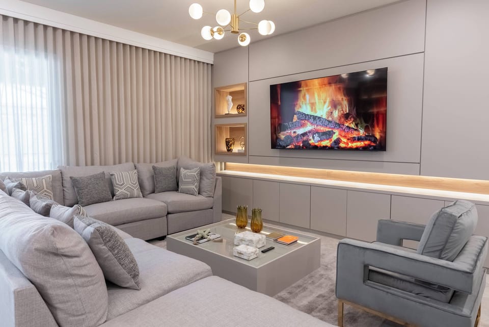 Welcome to our beautifully designed living room, featuring soft gray sofas perfect for relaxing by the cozy fireplace. Large windows with light curtains bring in natural light, enhancing the modern decor and creating a warm.