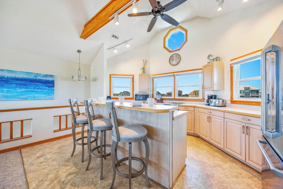 Gather around this sunlit kitchen island with vaulted ceilings and ocean-inspired touches. Natural light floods through every window, illuminating your morning coffee moments and meal prep under the warm glow of exposed beams.