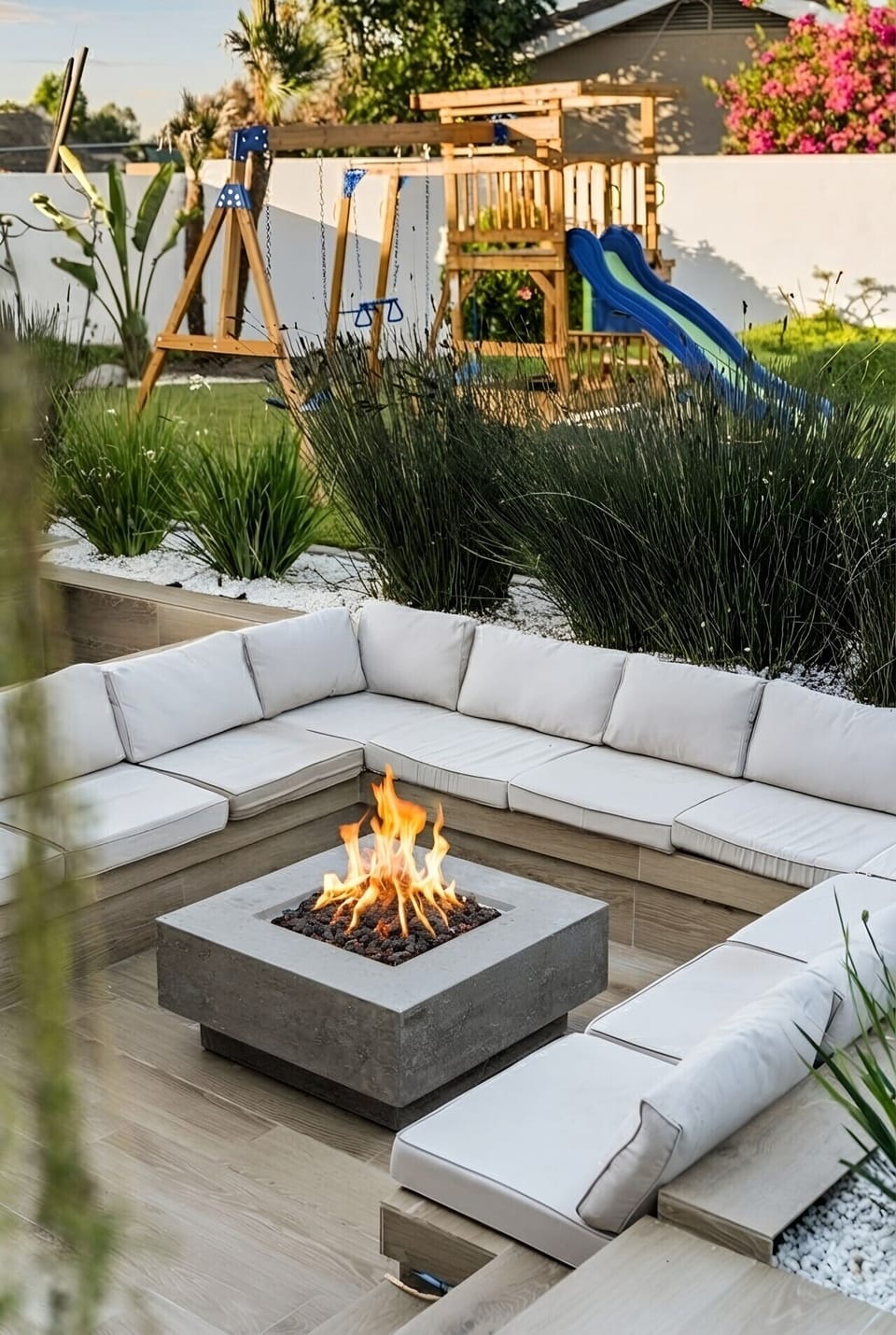 Sunken conversation area, plush cushions wrapping a crackling firepit, with a wooden play structure and lush landscaping just beyond. A refined spot for evening gatherings, while little ones enjoy their own corner of the yard.